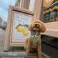 Dog wearing a floral harness and bow tie sitting in front of a lemon-themed sign and store window.