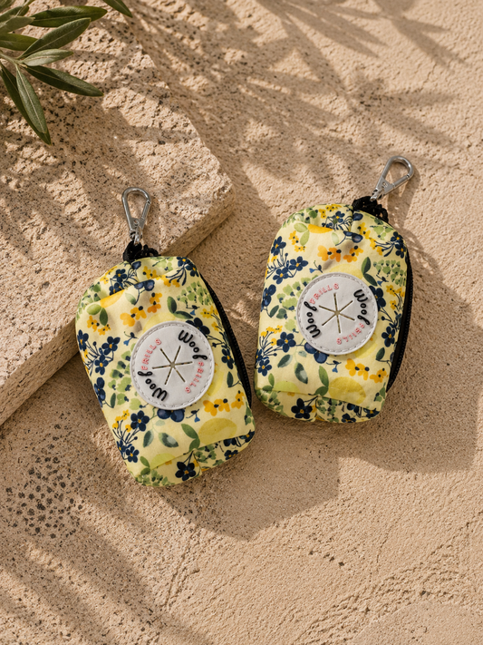 Two floral-patterned poo bag holders on a sandy surface with palm leaves.