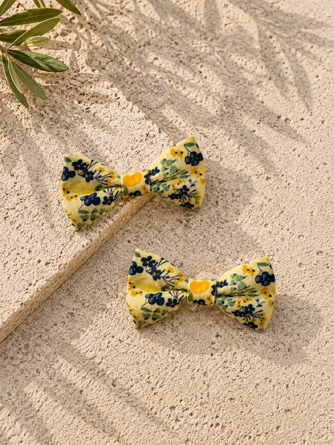 Two yellow floral dog bow ties on a textured beige surface with leaves in the corner.