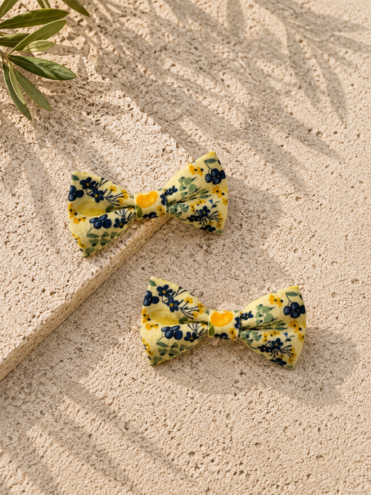 Two yellow floral dog bow ties on a textured beige surface with leaves in the corner.